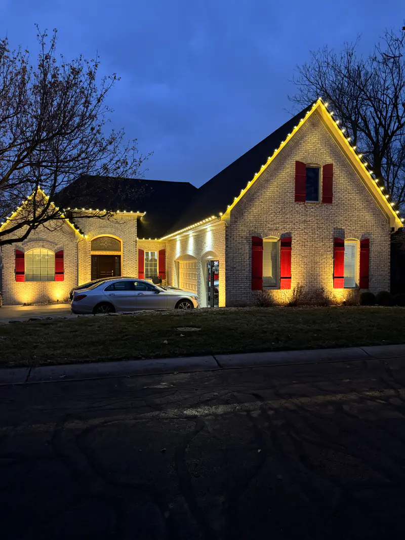 Light brick house at dusk with warm white bulbs tracing every gable and eave, red shutters glowing under uplights