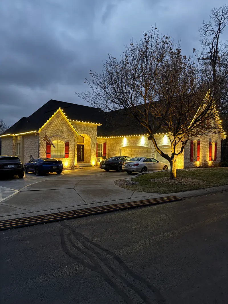 The same brick house from the street, warm white roofline lights above a lit bare tree and two parked cars