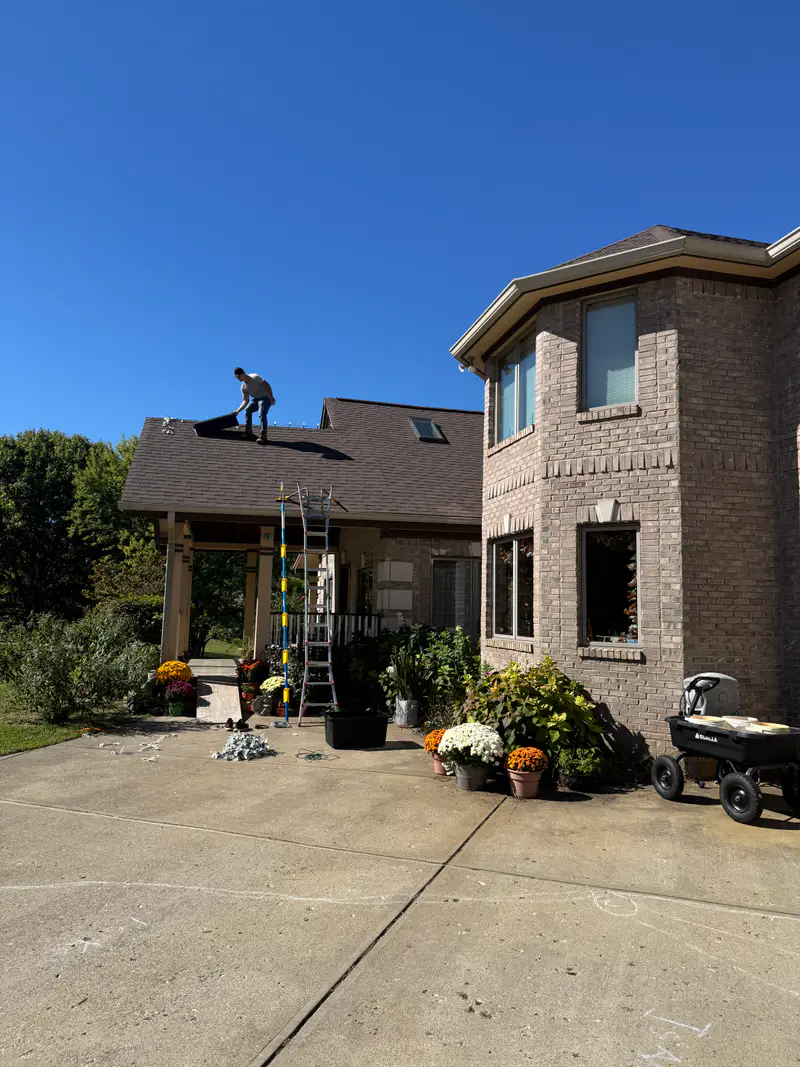 Installer on the roof of a brick home while fall mums line the driveway below