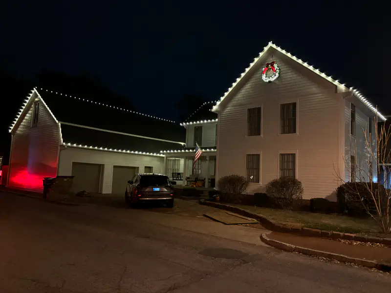 Two story white colonial at night outlined in warm white bulbs with a lit wreath centred in the front gable