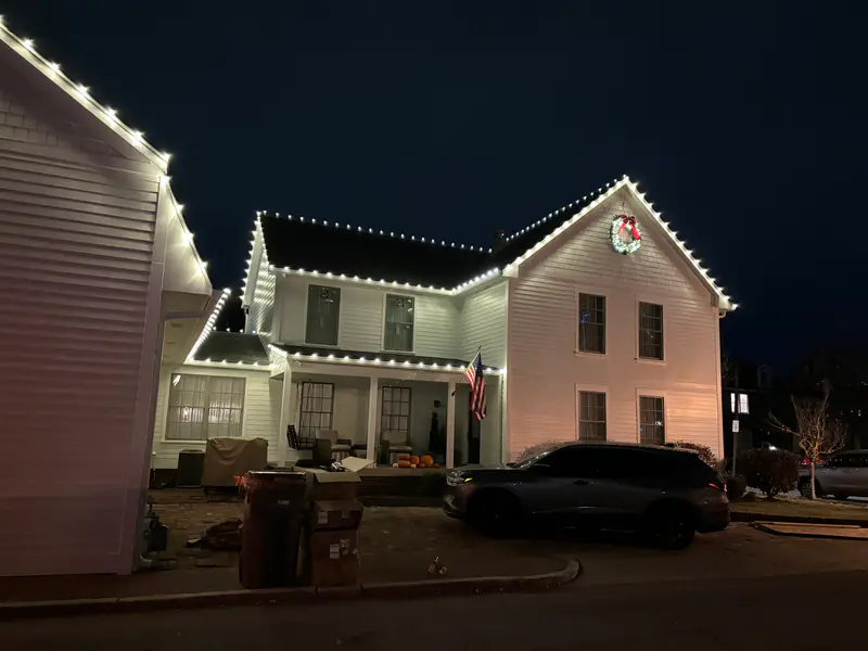 Side view of the white colonial, warm white lights running up both gables to the lit wreath