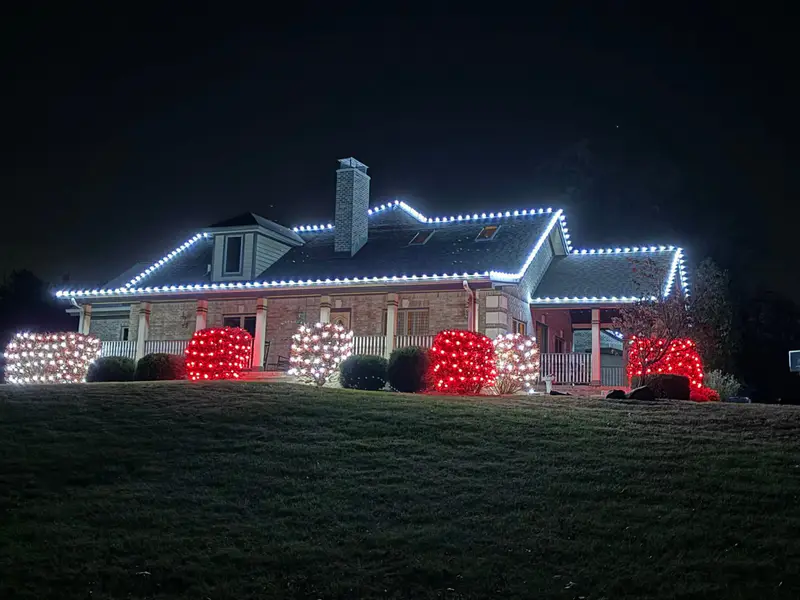 Brick house outlined in cool white bulbs with red and white lit shrubs across the front beds