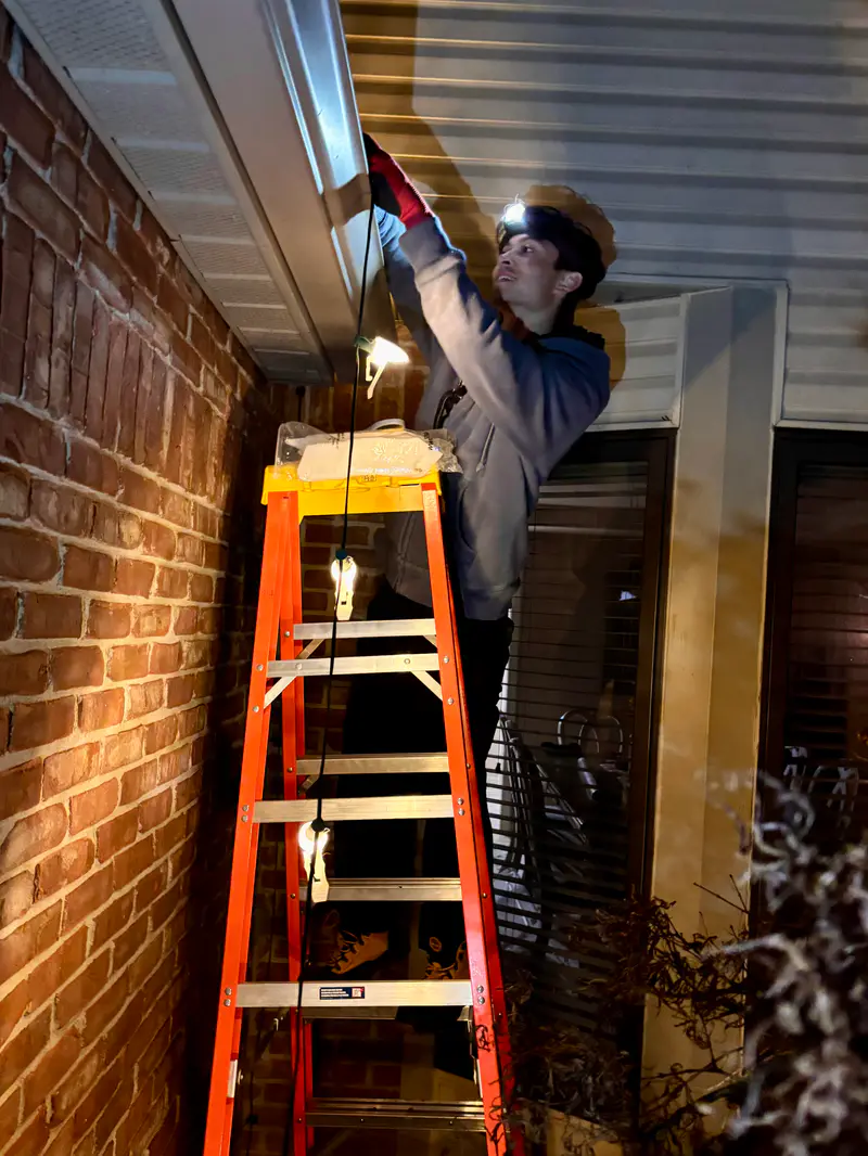 Installer on a ladder beside a brick wall at dusk, fitting bulbs along the eave by headlamp