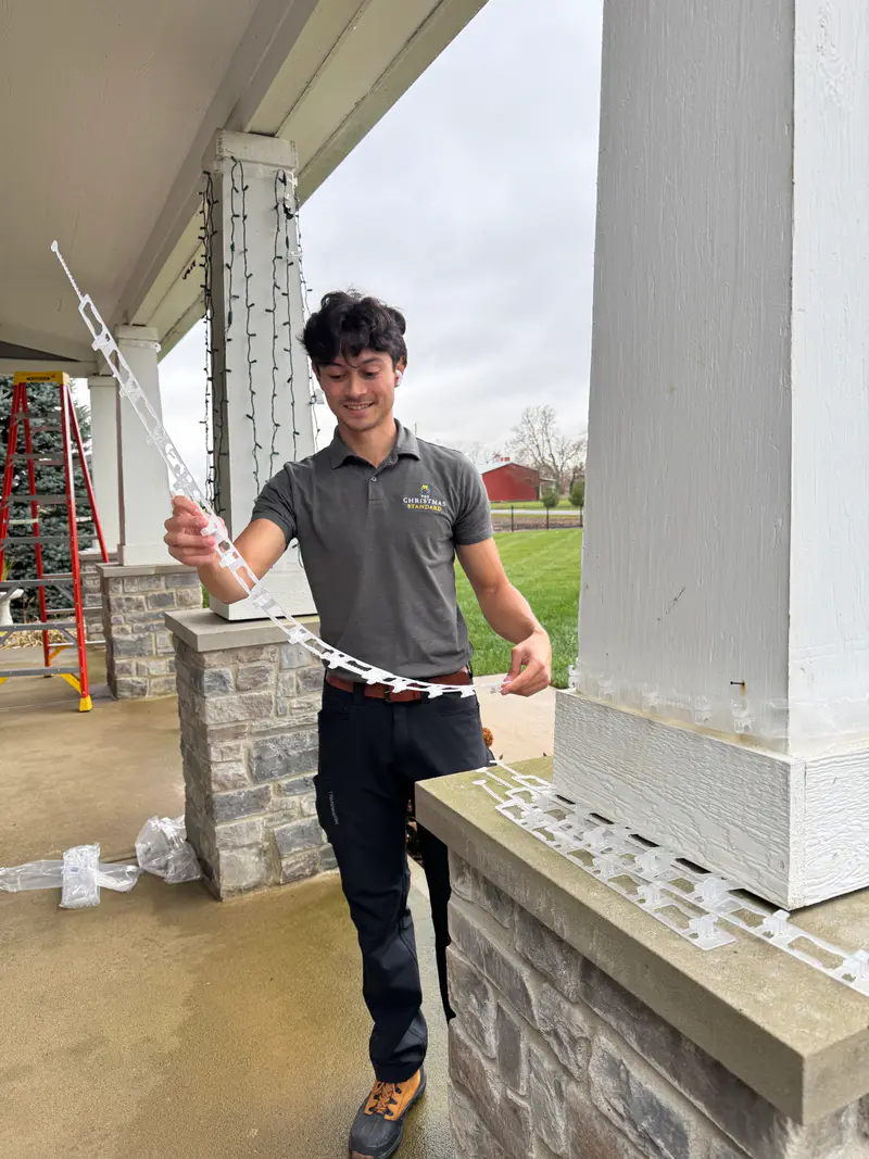 Kyle in a branded Christmas Standard shirt holding a run of bulbs beside a porch column