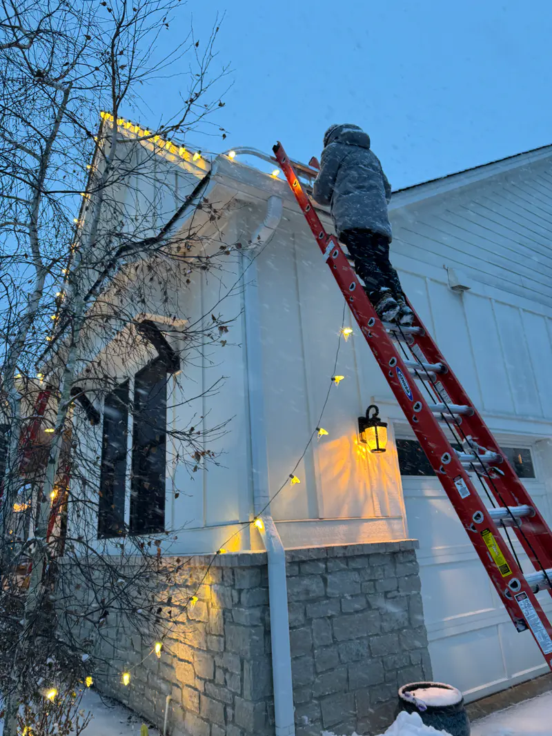 Installer on a tall ladder at dusk, the first lights of the roofline already on