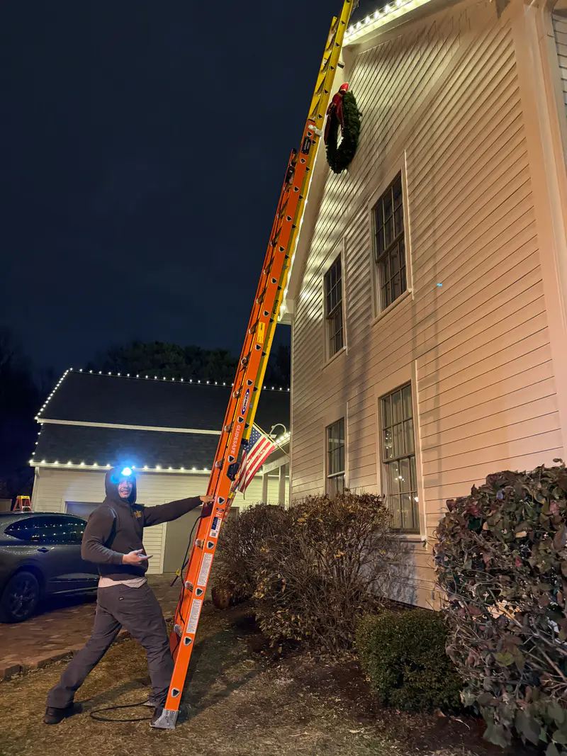 Installer with a headlamp at the foot of a tall ladder at night, a lit wreath and roofline above