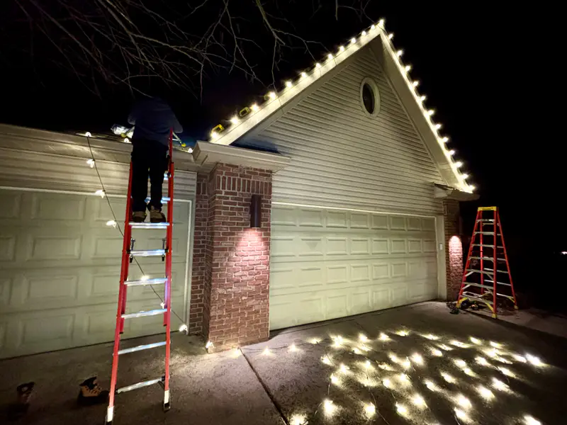 Installer on a ladder at night fitting warm white bulbs along a garage gable