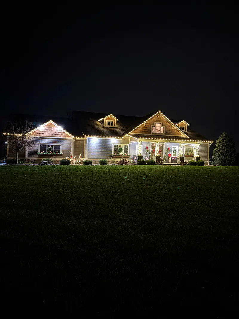 Wide ranch house across a dark lawn, warm and cool white lights along the roofline and gables