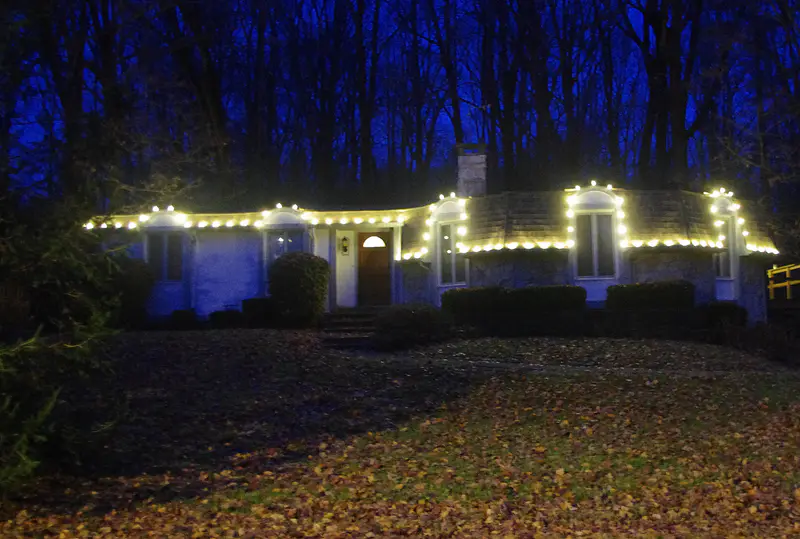 Long ranch house at blue hour with a single warm white line of bulbs stepping over every dormer, woods behind