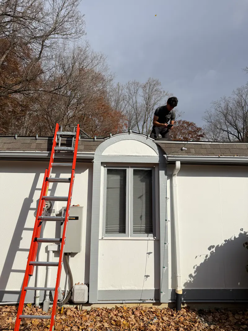 Installer working the roof edge above an arched window, a ladder against the white wall below