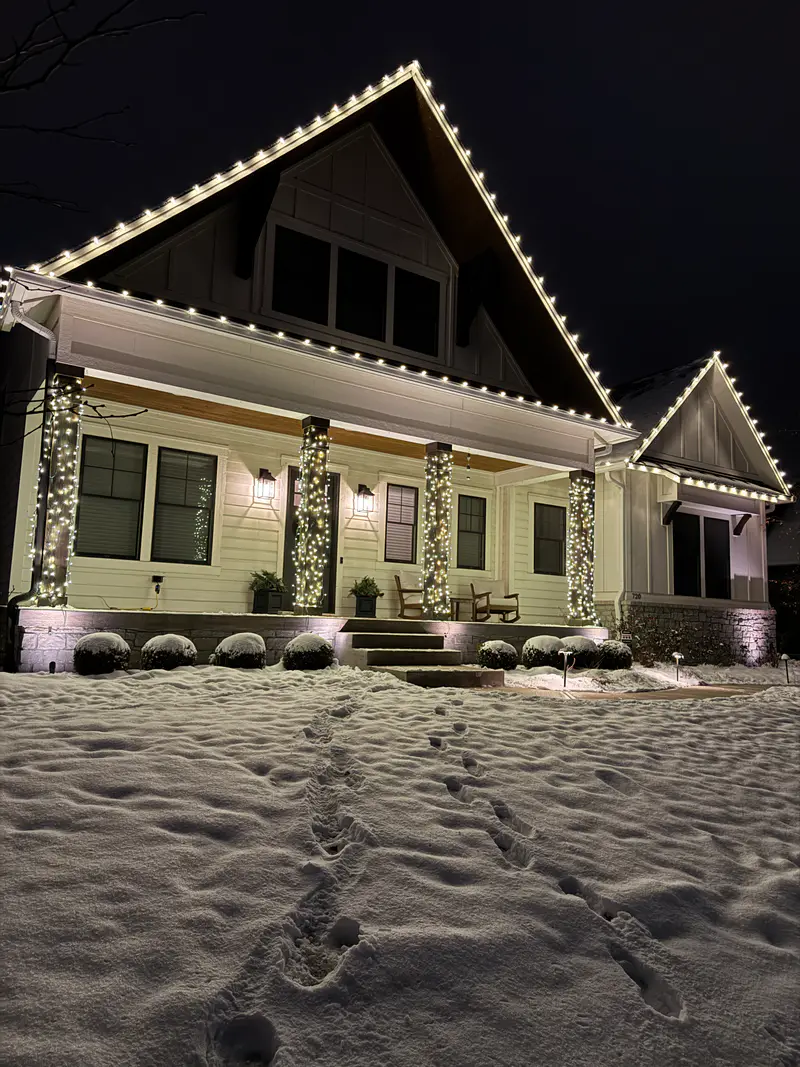 Craftsman porch in fresh snow with lit wrapped columns and warm white bulbs up the steep gable