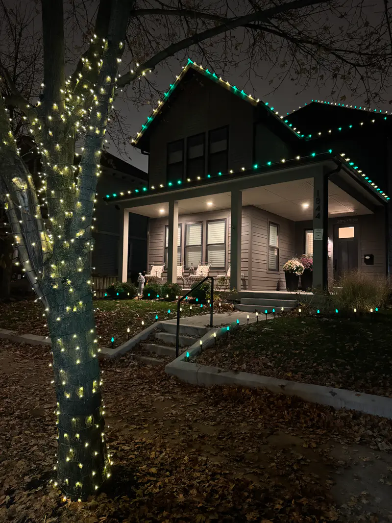 Tree trunk wrapped in warm lights in front of a porch lit with a green and white roofline
