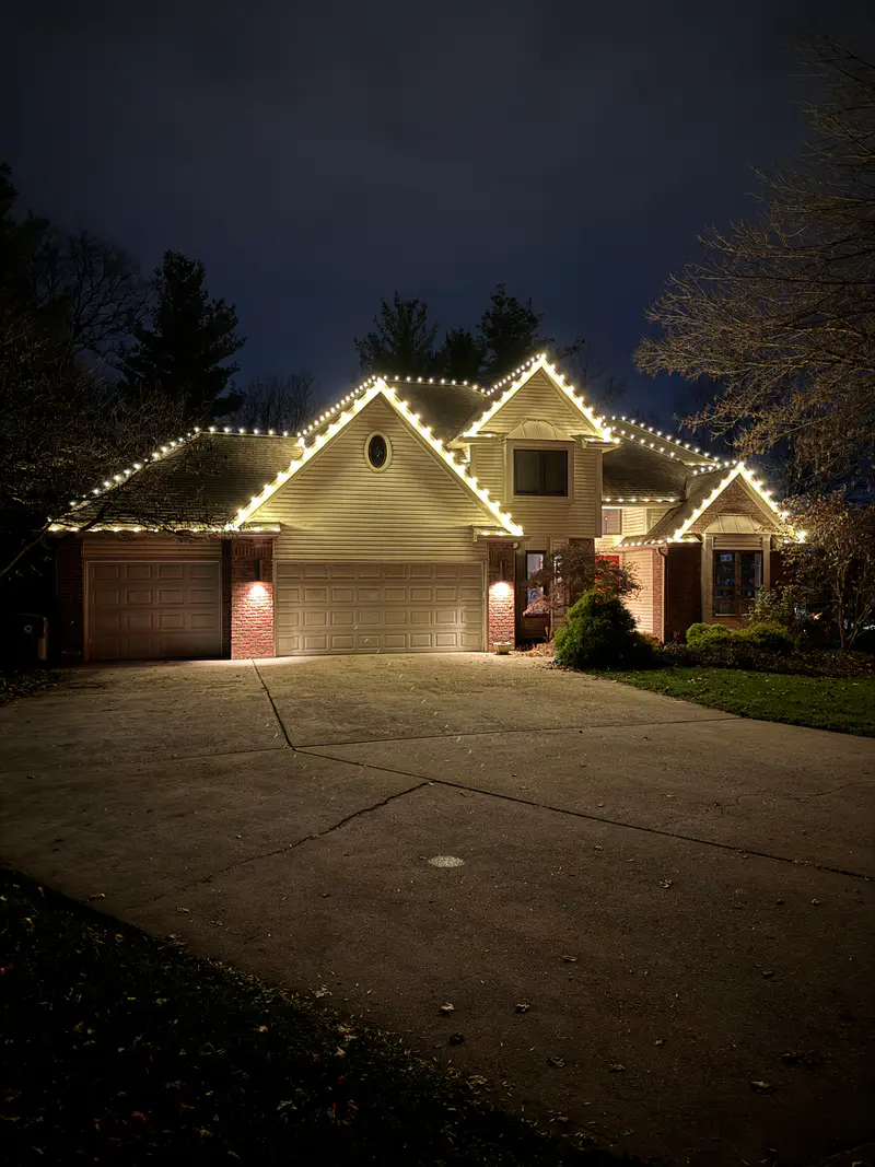 Two gabled house with warm white lights on every roof edge, seen from the end of a long driveway