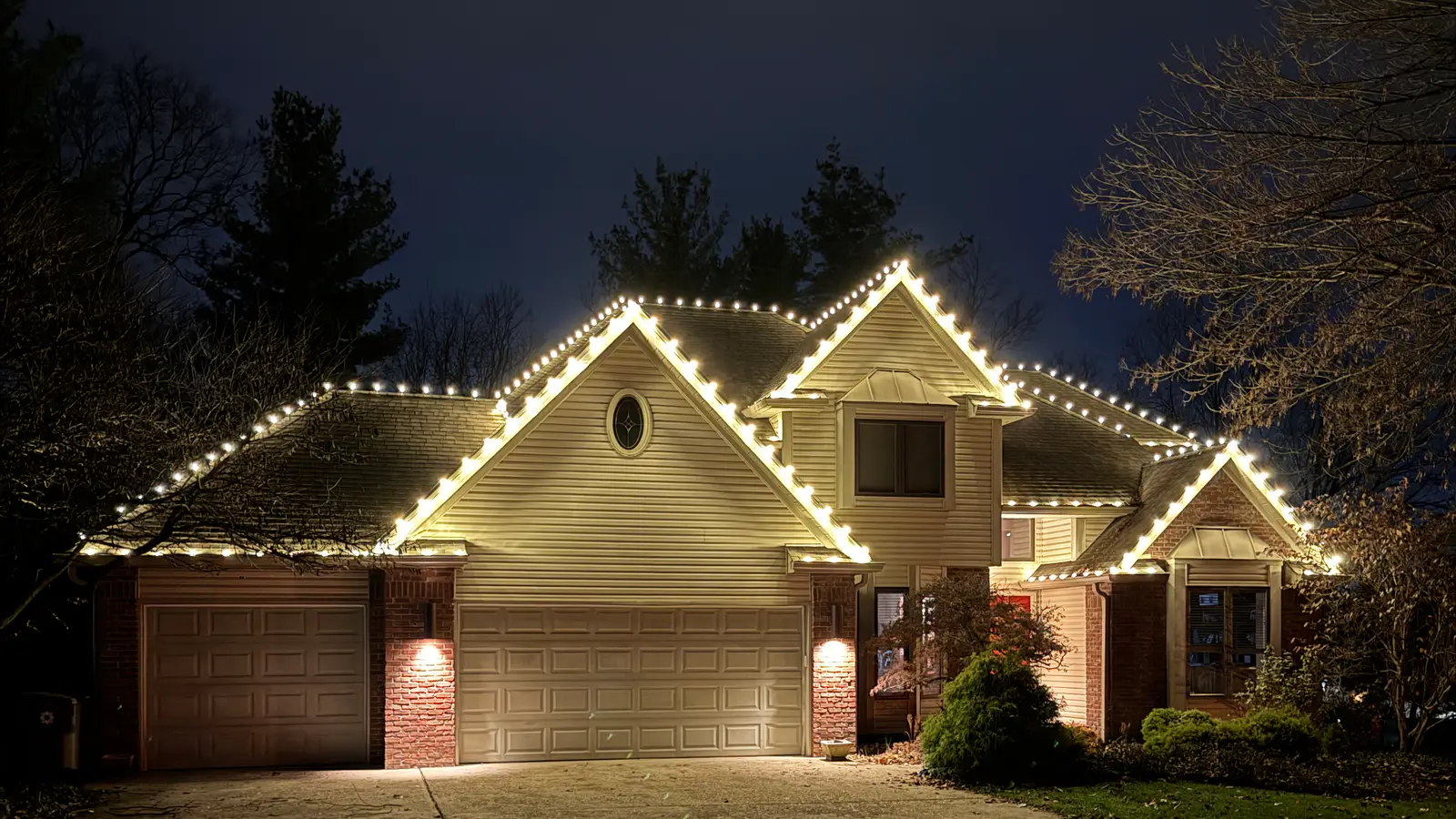 Two gabled house with warm white lights on every roof edge, seen from the end of a long driveway