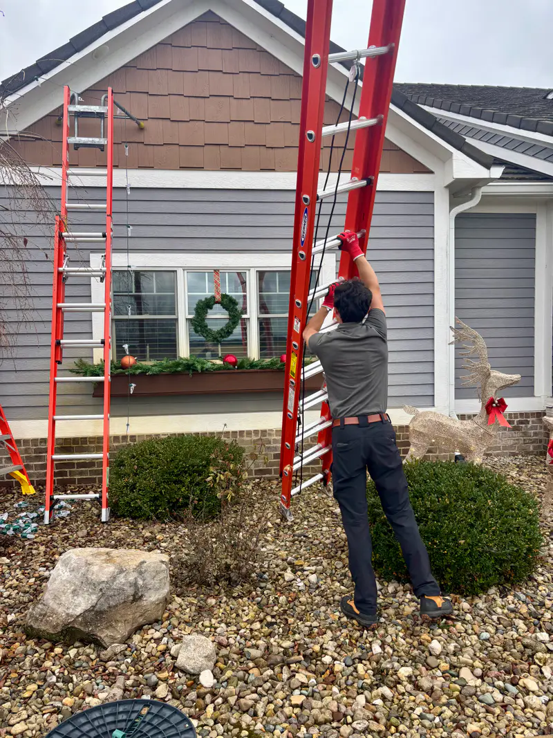 Installer on a ladder beside a window with a wreath, a second ladder set against the house